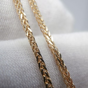 May include: Close-up of two gold-colored chain necklaces. The chains have a woven design, with each link interlocked to create a textured surface. The chains are set against a white background.