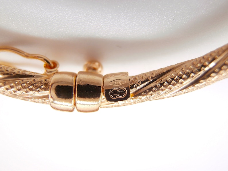 May include: Close-up of a gold bangle bracelet with a twisted design and a clasp. The bracelet is marked with "375" and a logo.