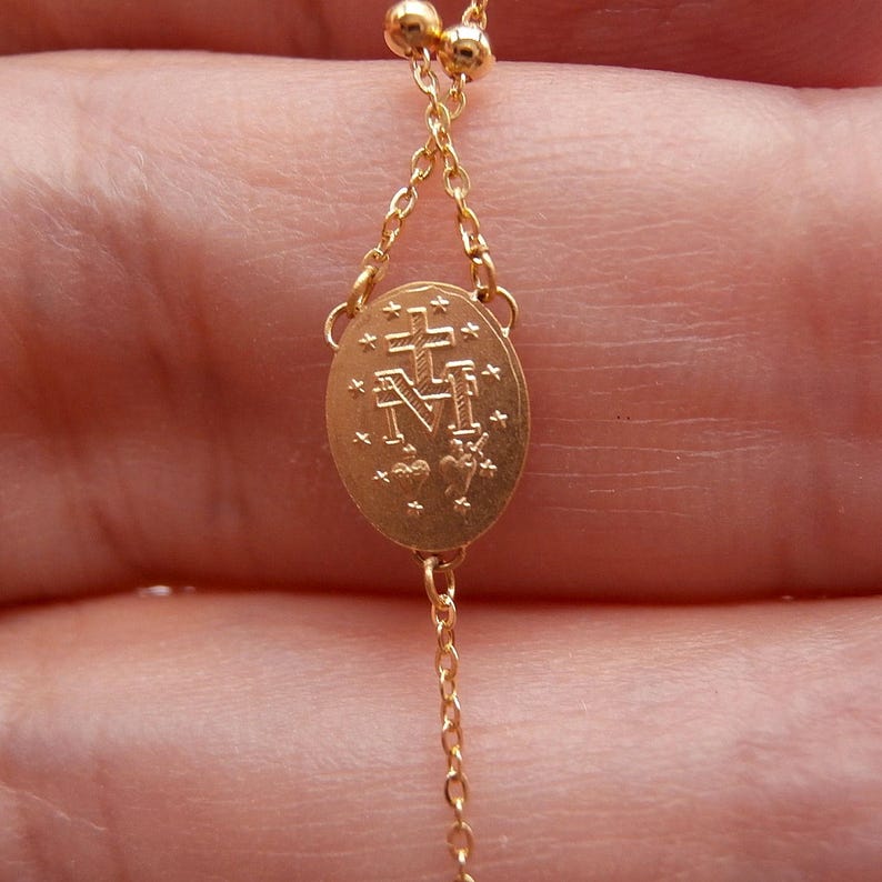 May include: Close-up of a gold-colored rosary chain. The chain features a central oval pendant with an engraved cross and the letter "M". The rosary is made of small, delicate links and beads.