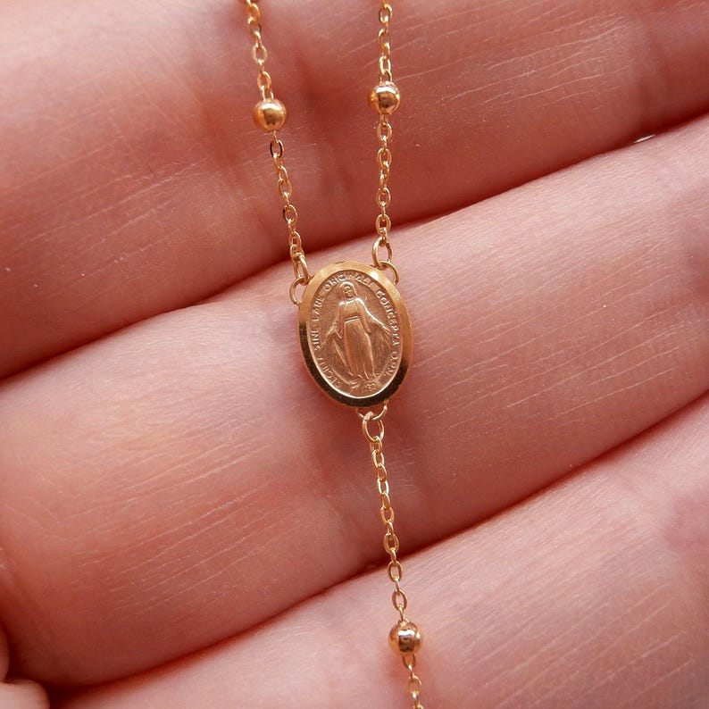 May include: A gold-colored rosary necklace with a detailed oval pendant featuring a religious figure. The necklace has small, round beads and delicate chain links. The rosary is displayed against a soft, blurred background.