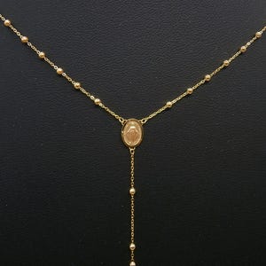May include: A gold-colored necklace featuring a central oval pendant with a religious figure. The necklace has a delicate chain with small, round beads and a longer chain with beads hanging below the pendant. The necklace is displayed against a dark background.