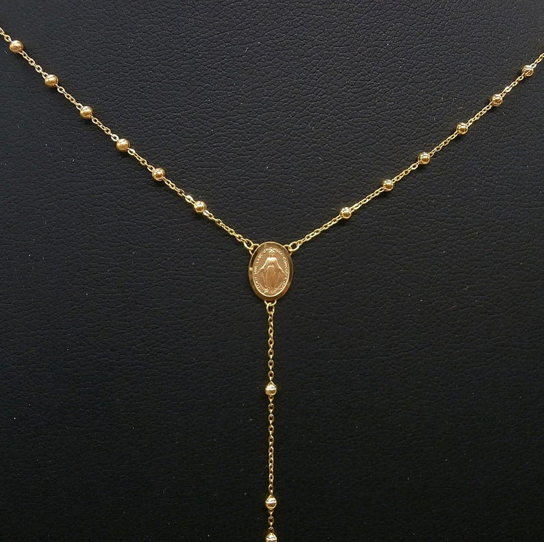 May include: A gold-colored necklace featuring a central oval pendant with a religious figure. The necklace has a delicate chain with small, round beads and a longer chain with beads hanging below the pendant. The necklace is displayed against a dark background.