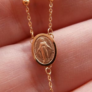 May include: Close-up of a gold-colored rosary chain. The chain features small, round beads and a pendant with an oval medallion. The medallion has an image of a figure and text around the edge.
