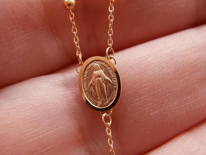 May include: Close-up of a gold-colored rosary chain. The chain features small, round beads and a pendant with an oval medallion. The medallion has an image of a figure and text around the edge.
