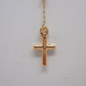 May include: A gold-colored cross pendant hanging from a delicate chain. The cross has a classic design with a vertical bar intersecting a horizontal bar. The chain and cross are made of a shiny, reflective metal.