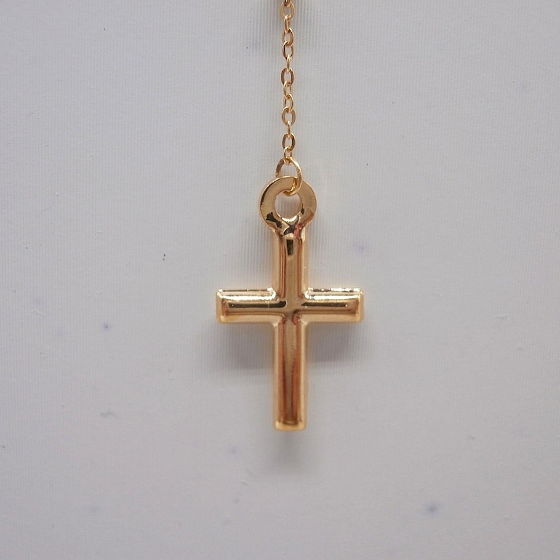May include: A gold-colored cross pendant hanging from a delicate chain. The cross has a classic design with a vertical bar intersecting a horizontal bar. The chain and cross are made of a shiny, reflective metal.