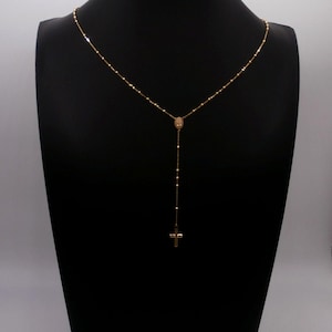 May include: A gold-colored rosary necklace with a cross pendant. The necklace is displayed on a black mannequin. The chain is delicate, with small beads and a larger centerpiece. The cross is a simple, classic design.