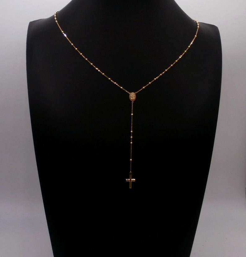 May include: A gold-colored rosary necklace with a cross pendant. The necklace is displayed on a black mannequin. The chain is delicate, with small beads and a larger centerpiece. The cross is a simple, classic design.