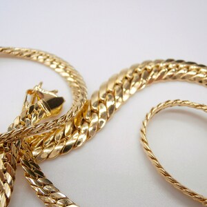 May include: Two gold chains, one with a thicker, more intricate design and the other with a thinner, more simple design. The chains are intertwined and laid out on a white surface.