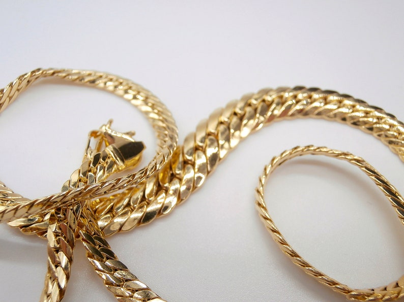 May include: Two gold chains, one with a thicker, more intricate design and the other with a thinner, more simple design. The chains are intertwined and laid out on a white surface.