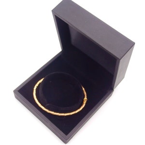 May include: A gold bangle bracelet in a black velvet lined jewelry box. The box is open and the bracelet is lying inside.