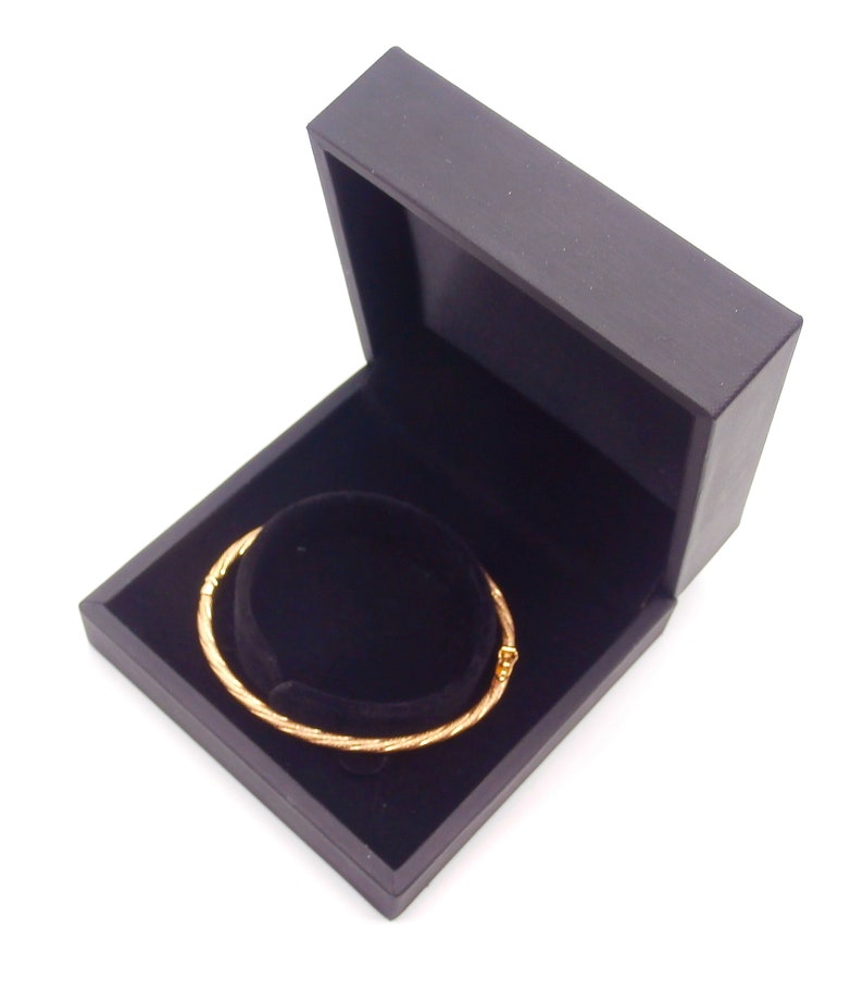 May include: A gold bangle bracelet in a black velvet lined jewelry box. The box is open and the bracelet is lying inside.