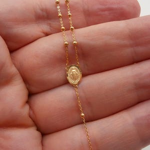 May include: A delicate gold-colored rosary necklace. The necklace features small, round beads and a central oval pendant with a religious figure. The chain is fine and elegant, suitable for religious or everyday wear.