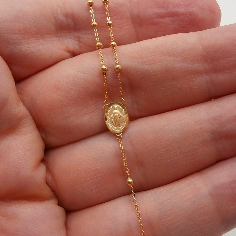 May include: A delicate gold-colored rosary necklace. The necklace features small, round beads and a central oval pendant with a religious figure. The chain is fine and elegant, suitable for religious or everyday wear.