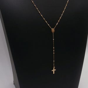 May include: A gold-colored necklace with a cross pendant. The necklace features a Y-shaped design with a small pendant at the top and a cross at the bottom. The chain is made of small beads.