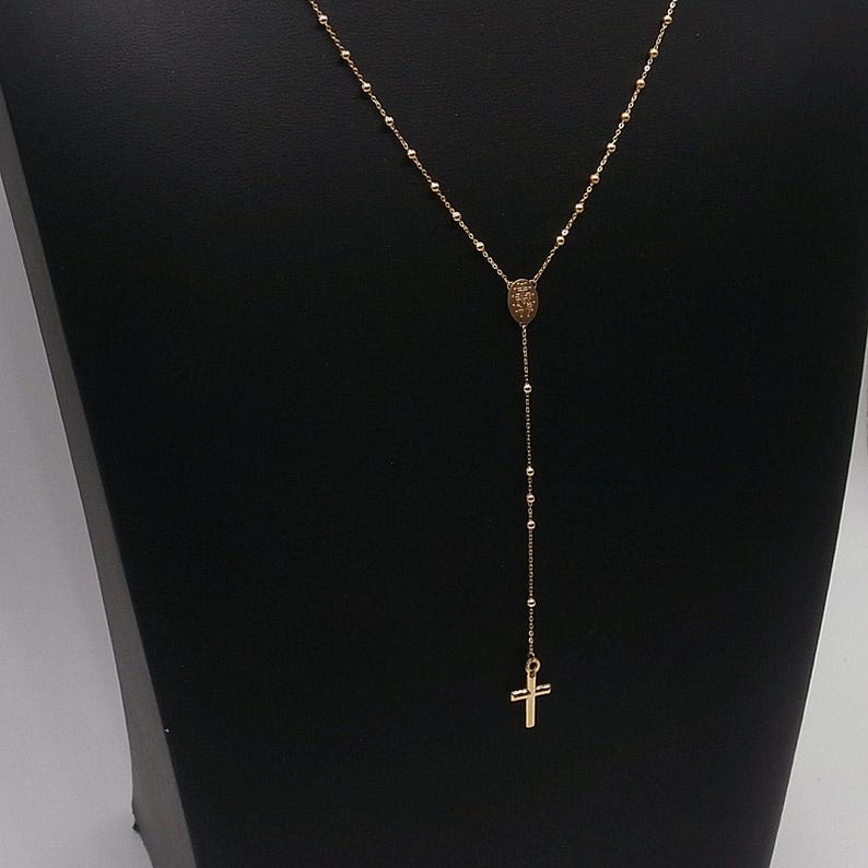 May include: A gold-colored necklace with a cross pendant. The necklace features a Y-shaped design with a small pendant at the top and a cross at the bottom. The chain is made of small beads.