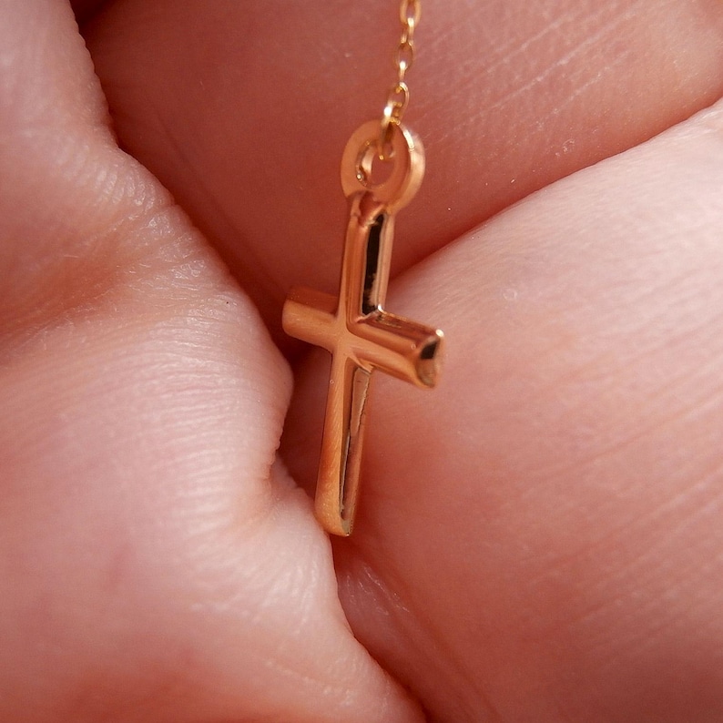 May include: A close-up of a gold-colored cross pendant. The cross is a simple, classic design with a polished finish. The pendant is suspended from a delicate chain, suggesting it is a necklace. The background is blurred, focusing attention on the jewelry.