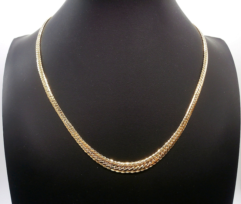 May include: A gold chain necklace with a flat, textured design. The chain is on a black display stand.
