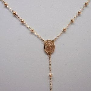 May include: Gold-colored rosary necklace featuring a central oval pendant with a religious figure. The necklace has a delicate chain with small, round beads and a longer chain with a few beads at the end.
