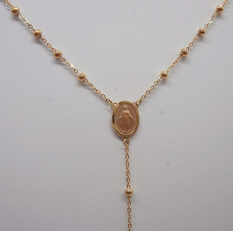 May include: Gold-colored rosary necklace featuring a central oval pendant with a religious figure. The necklace has a delicate chain with small, round beads and a longer chain with a few beads at the end.