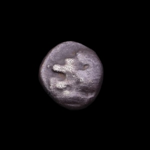 Puede incluir: Primer plano de una antigua moneda de plata sobre un fondo negro. La moneda tiene una forma irregular y una superficie texturizada. Un diseño es visible en la cara de la moneda, posiblemente un símbolo o inscripción.