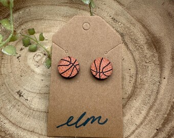 May include: A pair of round stud earrings featuring a basketball design. The earrings are orange with black lines, mounted on a brown tag with the handwritten word "elm". The earrings are displayed on a wooden surface.