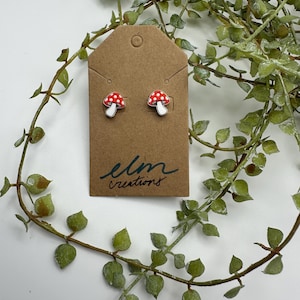 May include: A pair of mushroom stud earrings with red caps and white polka dots, displayed on a brown tag with the text "elm creations". The earrings are set against a backdrop of green foliage.