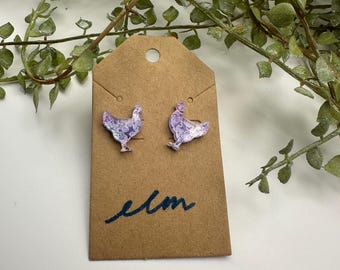 May include: A pair of stud earrings featuring a chicken design with a floral pattern in shades of purple and white. The earrings are displayed on a brown tag with the handwritten word "elm" in blue ink.