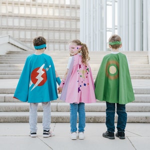May include: Three children wearing superhero capes stand on a set of stairs. The child in the middle is wearing a pink cape with stars, the child on the left is wearing a blue cape with a lightning bolt, and the child on the right is wearing a green cape with a circle.