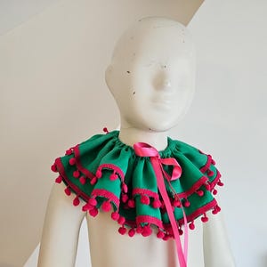 K&ouml;nnte beinhalten: Ein gr&uuml;ner R&uuml;schenkragen mit rosa Besatz und Bommeln, gebunden mit einem rosa Band. Der Kragen wird auf einer wei&szlig;en Schaufensterpuppe pr&auml;sentiert, die sein festliches Design zeigt. Dieser dekorative Kragen eignet sich f&uuml;r Kost&uuml;me oder besondere Anl&auml;sse.