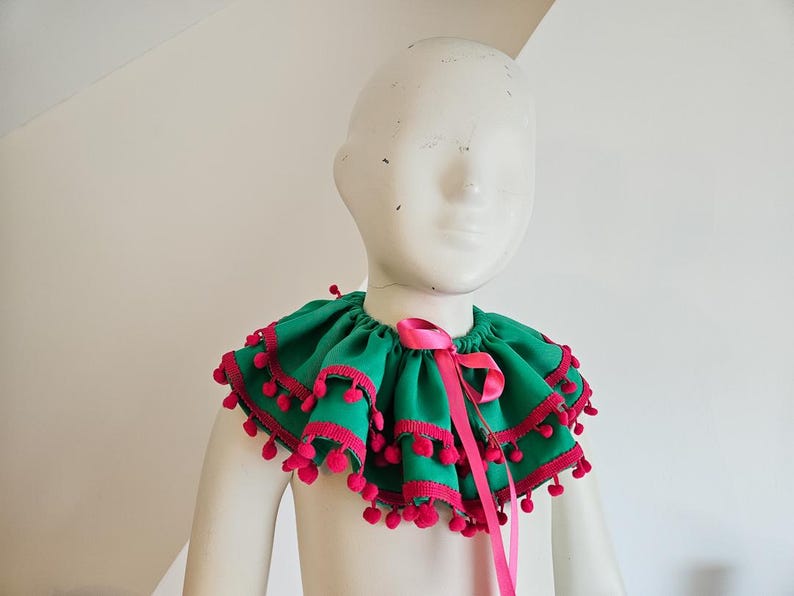 K&ouml;nnte beinhalten: Ein gr&uuml;ner R&uuml;schenkragen mit rosa Besatz und Bommeln, gebunden mit einem rosa Band. Der Kragen wird auf einer wei&szlig;en Schaufensterpuppe pr&auml;sentiert, die sein festliches Design zeigt. Dieser dekorative Kragen eignet sich f&uuml;r Kost&uuml;me oder besondere Anl&auml;sse.