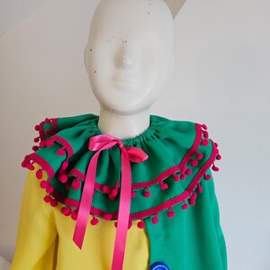 K&ouml;nnte beinhalten: Ein farbenfrohes Kost&uuml;m mit einem gr&uuml;n-gelben Oberteil und einem gro&szlig;en, ger&uuml;schten Kragen. Der Kragen ist mit rosa Pompons und einem rosa Band verziert. Ein gro&szlig;er blauer Knopf akzentuiert die gr&uuml;ne Seite des Kleidungsst&uuml;cks.