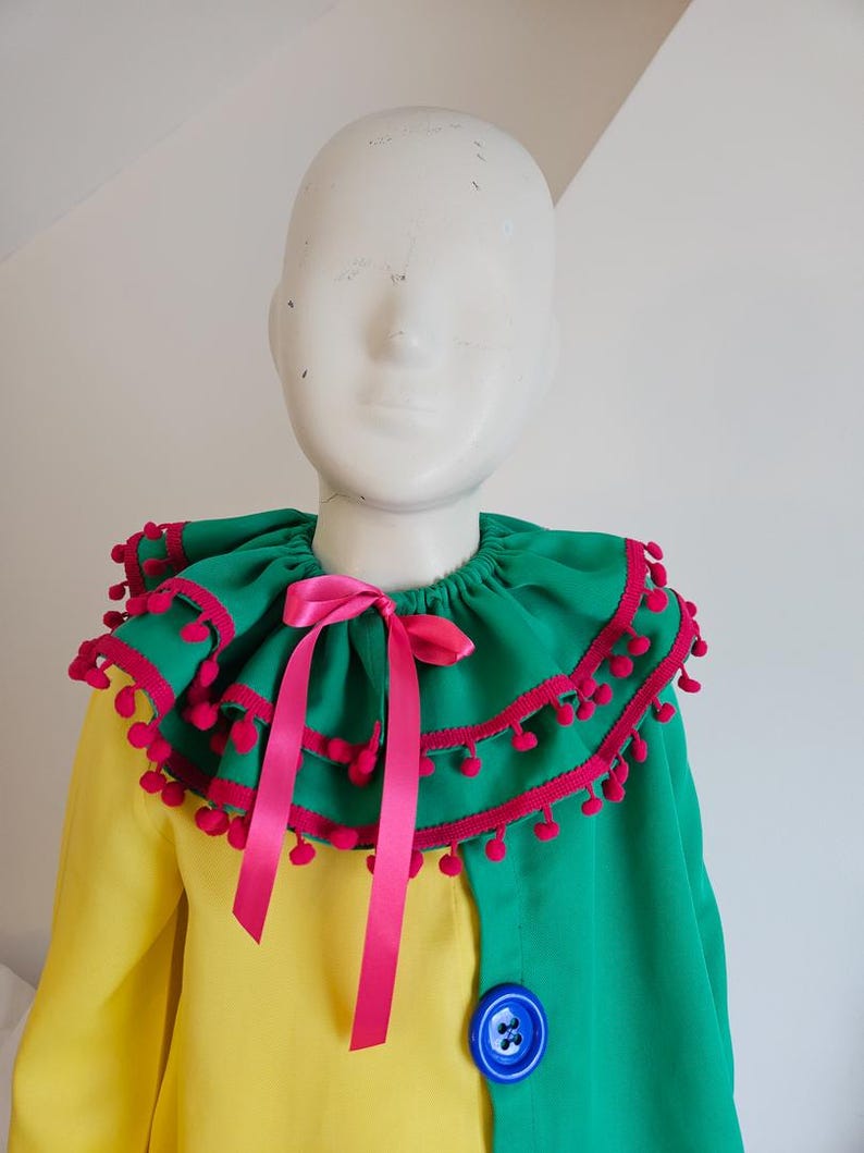 K&ouml;nnte beinhalten: Ein farbenfrohes Kost&uuml;m mit einem gr&uuml;n-gelben Oberteil und einem gro&szlig;en, ger&uuml;schten Kragen. Der Kragen ist mit rosa Pompons und einem rosa Band verziert. Ein gro&szlig;er blauer Knopf akzentuiert die gr&uuml;ne Seite des Kleidungsst&uuml;cks.