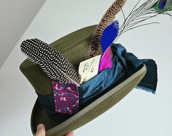 May include: A green top hat with a teal blue ribbon, pink feathers, a brown and white speckled feather, a blue feather, and a peacock feather. A small white tag with the text "10/6" is attached to the hat.