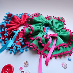 K&ouml;nnte beinhalten: Zwei farbenfrohe Haargummis, eines rot mit blauen Verzierungen und Bommeln, das andere gr&uuml;n mit rosa Verzierungen und Bommeln. Beide haben passende Schleifenb&auml;nder. Verschiedene Kn&ouml;pfe und Verzierungen sind verstreut.