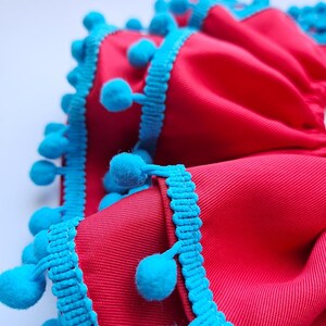 K&ouml;nnte beinhalten: Nahaufnahme eines leuchtend roten Stoffes mit strukturierter Oberfl&auml;che. Die Kanten sind mit leuchtend t&uuml;rkisfarbenen Bommelborten verziert. Der Stoff ist gefaltet, wodurch ein geschichteter Effekt entsteht, und die Bommel verleihen eine verspielte Note.