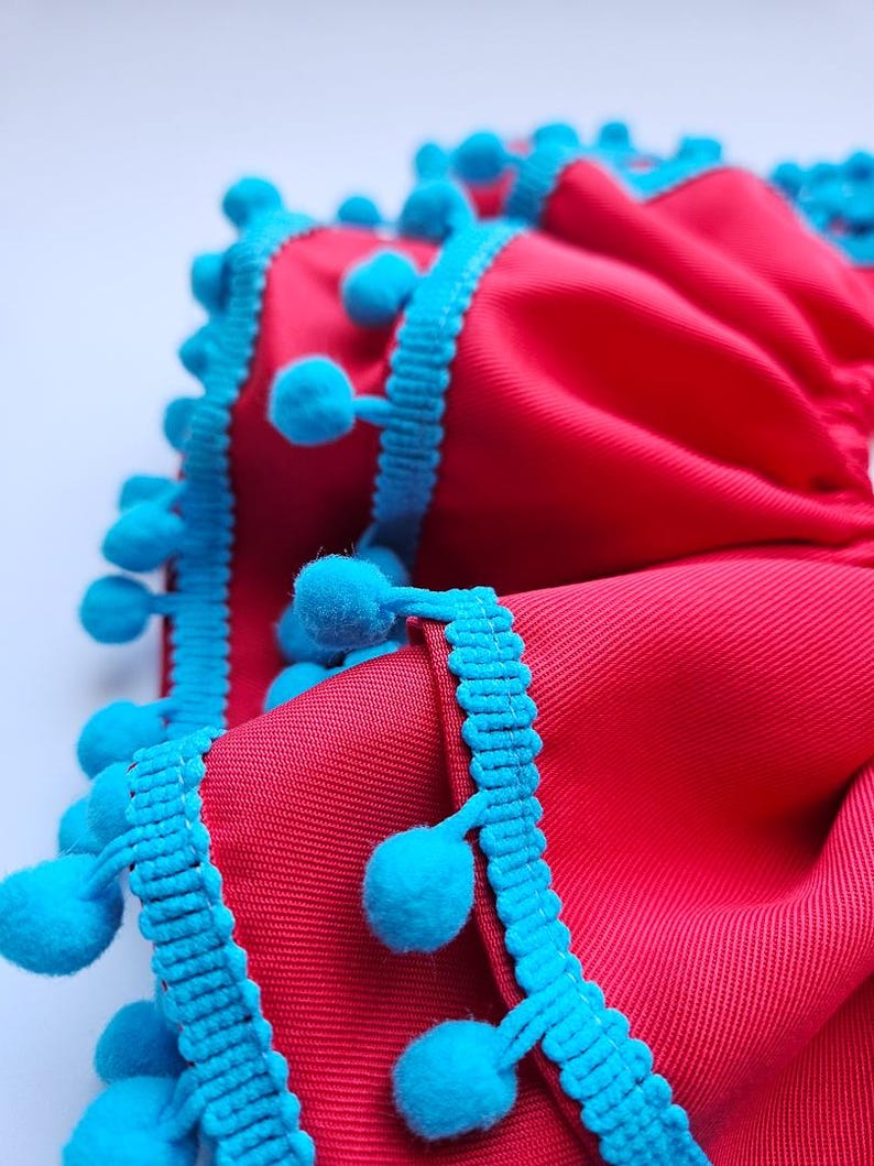 K&ouml;nnte beinhalten: Nahaufnahme eines leuchtend roten Stoffes mit strukturierter Oberfl&auml;che. Die Kanten sind mit leuchtend t&uuml;rkisfarbenen Bommelborten verziert. Der Stoff ist gefaltet, wodurch ein geschichteter Effekt entsteht, und die Bommel verleihen eine verspielte Note.