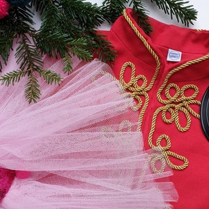 Può includere: Un costume rosso e oro con un cappello nero. Il costume ha un bordo in corda dorata e una gonna di tulle bianco. Il costume è per un bambino.