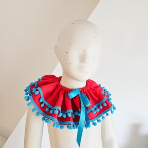 K&ouml;nnte beinhalten: Ein roter R&uuml;schenkragen mit blauer Bord&uuml;re und Bommeln. Vorne ist eine blaue Schleife gebunden. Der Kragen wird auf einer Schaufensterpuppe pr&auml;sentiert.