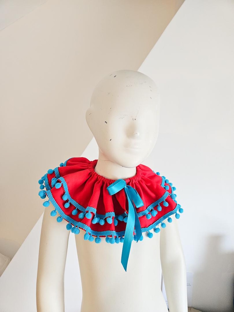 K&ouml;nnte beinhalten: Ein roter R&uuml;schenkragen mit blauer Bord&uuml;re und Bommeln. Vorne ist eine blaue Schleife gebunden. Der Kragen wird auf einer Schaufensterpuppe pr&auml;sentiert.