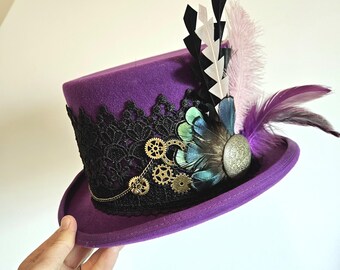 Può includere: Un cappello a cilindro viola con bordatura in pizzo nero, ingranaggi dorati e piume in tonalità di nero, bianco e viola.