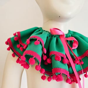 K&ouml;nnte beinhalten: Ein gr&uuml;ner R&uuml;schenkragen mit rosa Besatz und Bommeln. Ein rosa Satinband ist vorne zu einer Schleife gebunden. Der Kragen wird auf einer Schaufensterpuppe pr&auml;sentiert.