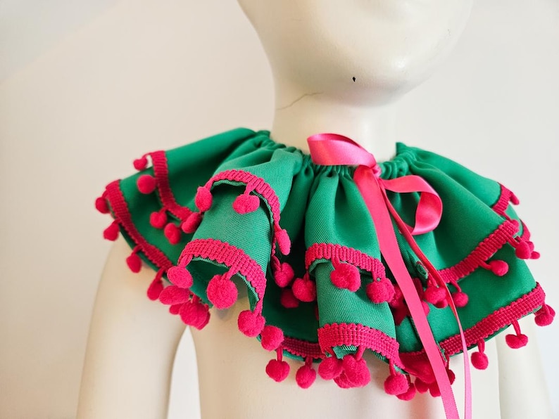 K&ouml;nnte beinhalten: Ein gr&uuml;ner R&uuml;schenkragen mit rosa Besatz und Bommeln. Ein rosa Satinband ist vorne zu einer Schleife gebunden. Der Kragen wird auf einer Schaufensterpuppe pr&auml;sentiert.