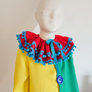 K&ouml;nnte beinhalten: Ein farbenfrohes Clownkost&uuml;m mit einem roten R&uuml;schenkragen mit blauen Pompons und einer passenden Schleife. Das Kost&uuml;m ist gelb und gr&uuml;n blockfarben gestaltet und mit blauen Kn&ouml;pfen versehen. Auf einer Schaufensterpuppe pr&auml;sentiert.