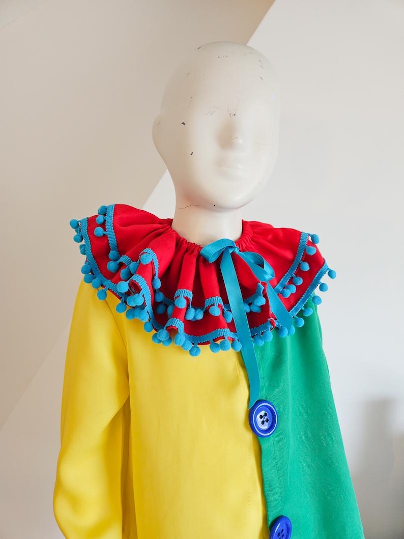 K&ouml;nnte beinhalten: Ein farbenfrohes Clownkost&uuml;m mit einem roten R&uuml;schenkragen mit blauen Pompons und einer passenden Schleife. Das Kost&uuml;m ist gelb und gr&uuml;n blockfarben gestaltet und mit blauen Kn&ouml;pfen versehen. Auf einer Schaufensterpuppe pr&auml;sentiert.