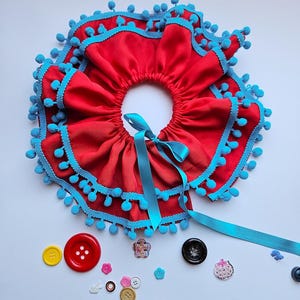 K&ouml;nnte beinhalten: Ein roter Stoffkragen mit R&uuml;schendesign, verziert mit blauen Pompons und B&auml;ndern. Verschiedene bunte Kn&ouml;pfe sind um den Kragen herum verstreut. Der Kragen ist zum Tragen um den Hals konzipiert.