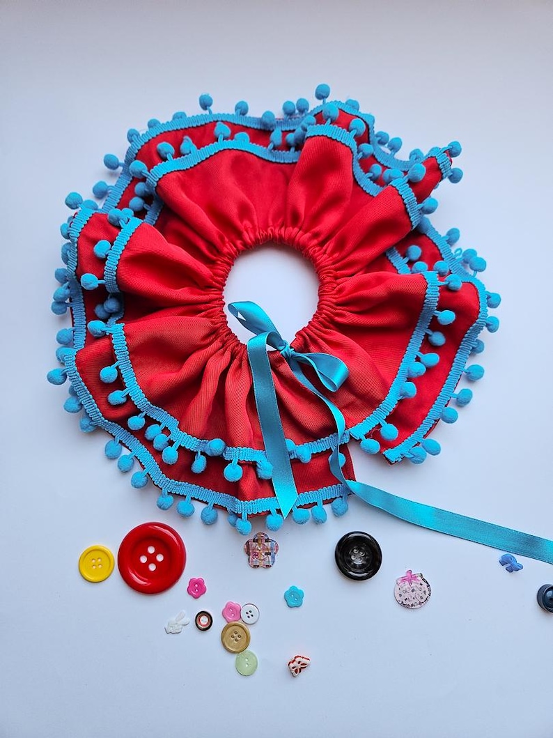 K&ouml;nnte beinhalten: Ein roter Stoffkragen mit R&uuml;schendesign, verziert mit blauen Pompons und B&auml;ndern. Verschiedene bunte Kn&ouml;pfe sind um den Kragen herum verstreut. Der Kragen ist zum Tragen um den Hals konzipiert.