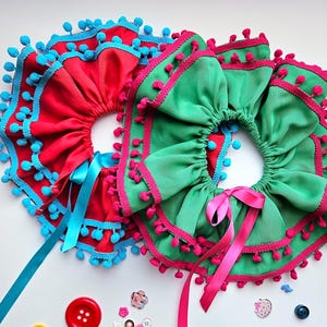 K&ouml;nnte beinhalten: Zwei dekorative R&uuml;schenkragen. Einer ist rot mit blauen Verzierungen und Bommeln, der andere ist gr&uuml;n mit rosa Verzierungen und Bommeln. Beide haben Schleifenb&auml;nder. Kn&ouml;pfe und Verzierungen sind verstreut.