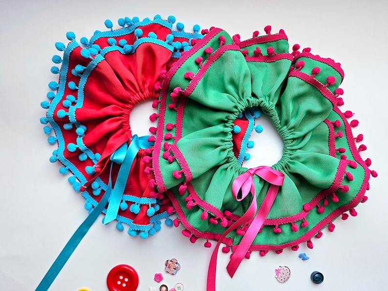 K&ouml;nnte beinhalten: Zwei dekorative R&uuml;schenkragen. Einer ist rot mit blauen Verzierungen und Bommeln, der andere ist gr&uuml;n mit rosa Verzierungen und Bommeln. Beide haben Schleifenb&auml;nder. Kn&ouml;pfe und Verzierungen sind verstreut.