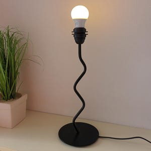 Op de afbeelding: Een zwarte tafellamp met een golvende steel en een ronde voet. De lamp heeft een witte LED-lamp.